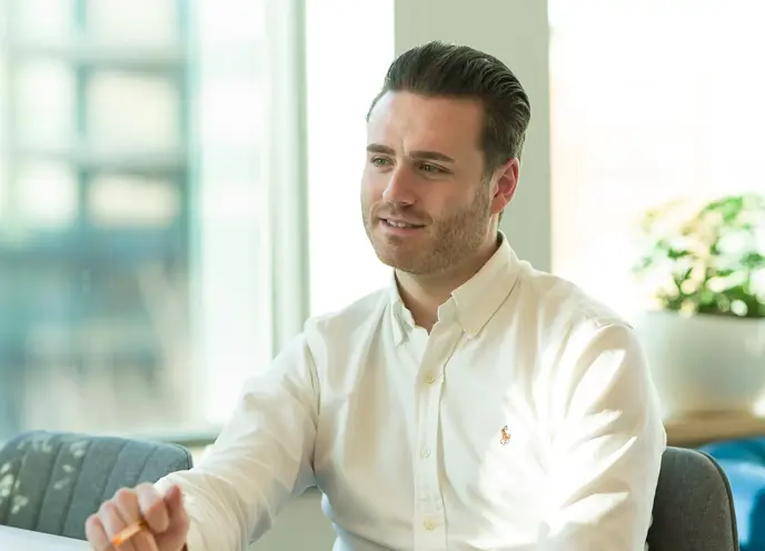 A young male in a white shirt sat at a desk holding a pen over a notebook with a confused expression.